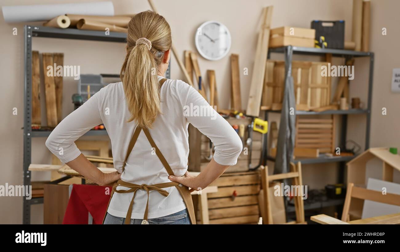 Blonde Woman In Workshop With Apron Facing Shelves Of Wood And Tools blonde-woman-in-workshop-with-apron-facing-shelves-of-wood-and-tools