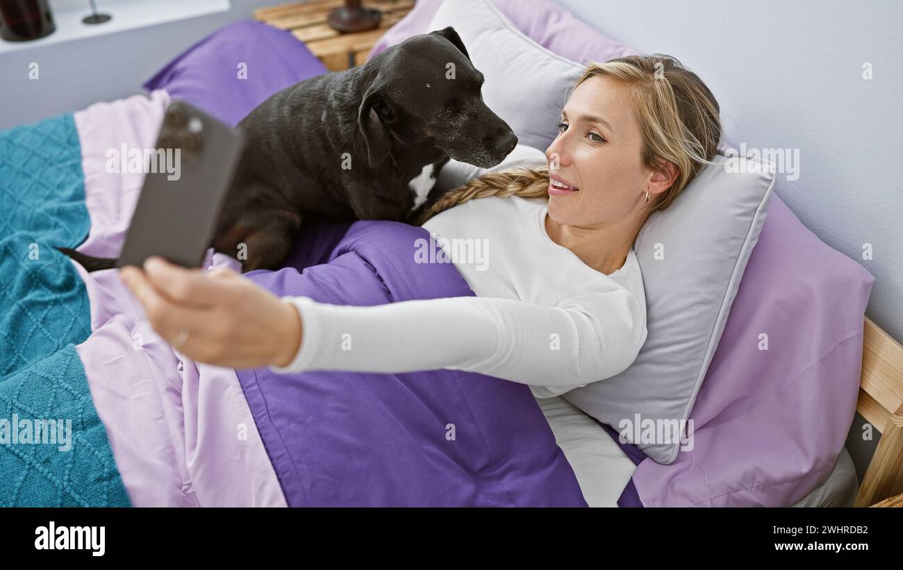 A young woman takes a selfie with her black labrador in a cozy bedroom ...