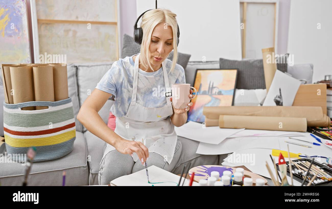 Beautiful blonde artist imbibing coffee and sound, a young woman's interior dance at the easel, drawing while listening to music in art studio Stock Photo