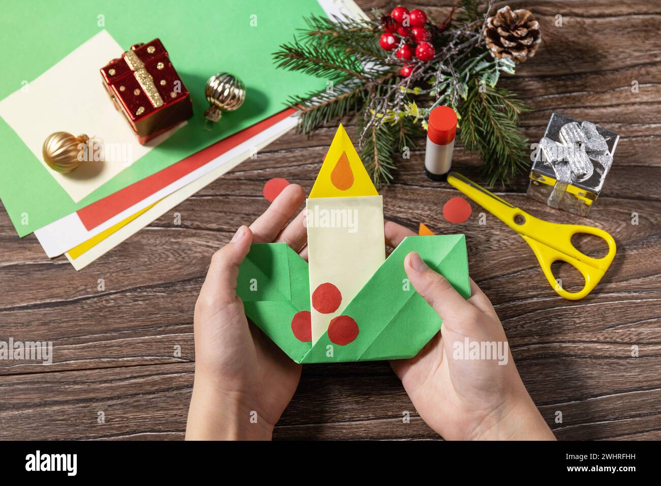 In the hands of a child Christmas greeting card gift origami candle on wooden table. Childrens art project, handmade, crafts for Stock Photo