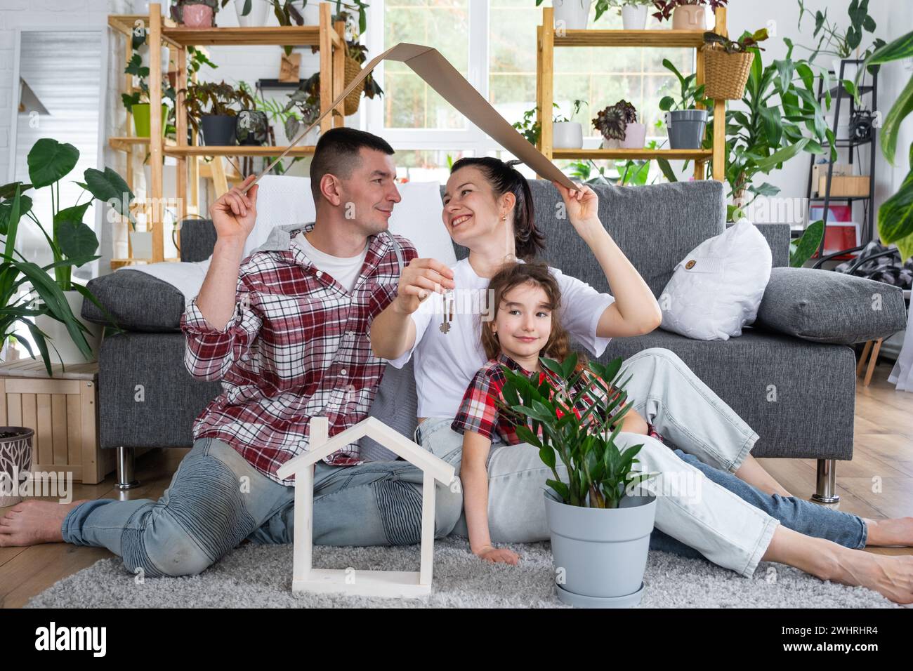 Traditional family with child are enjoying new home, sitting on the sofa and holding the roof. Mortgage, insurance and protectio Stock Photo