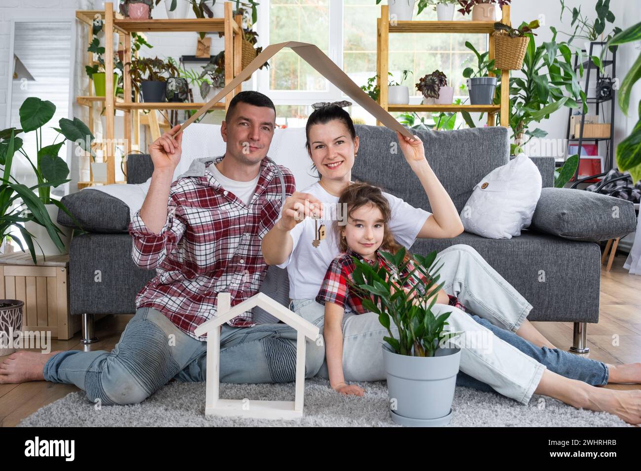 Traditional family with child are enjoying new home, sitting on the sofa and holding the roof. Mortgage, insurance and protectio Stock Photo