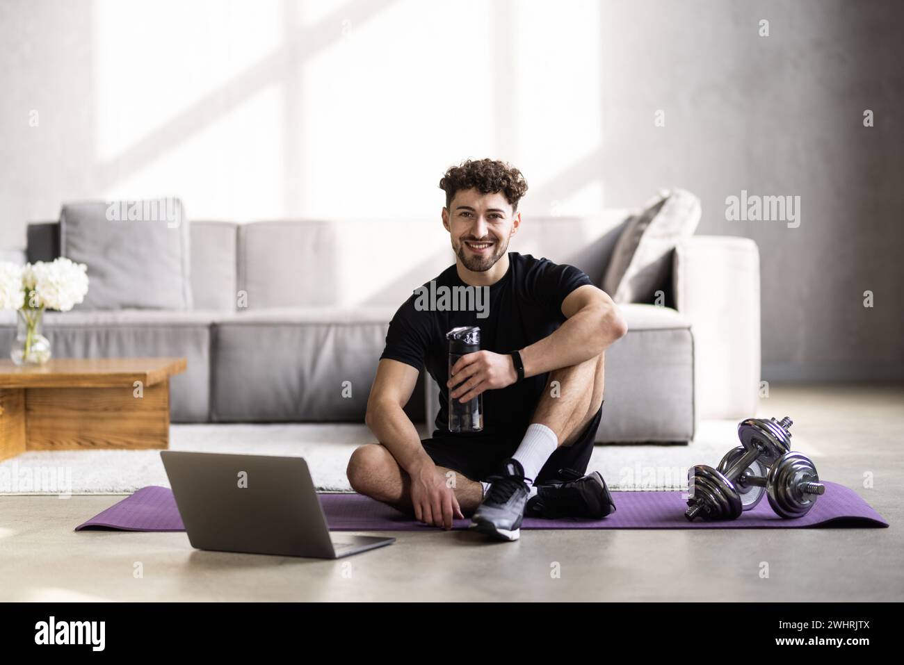 Smiling man using laptop and drinking water while having break during workout at home Stock Photo