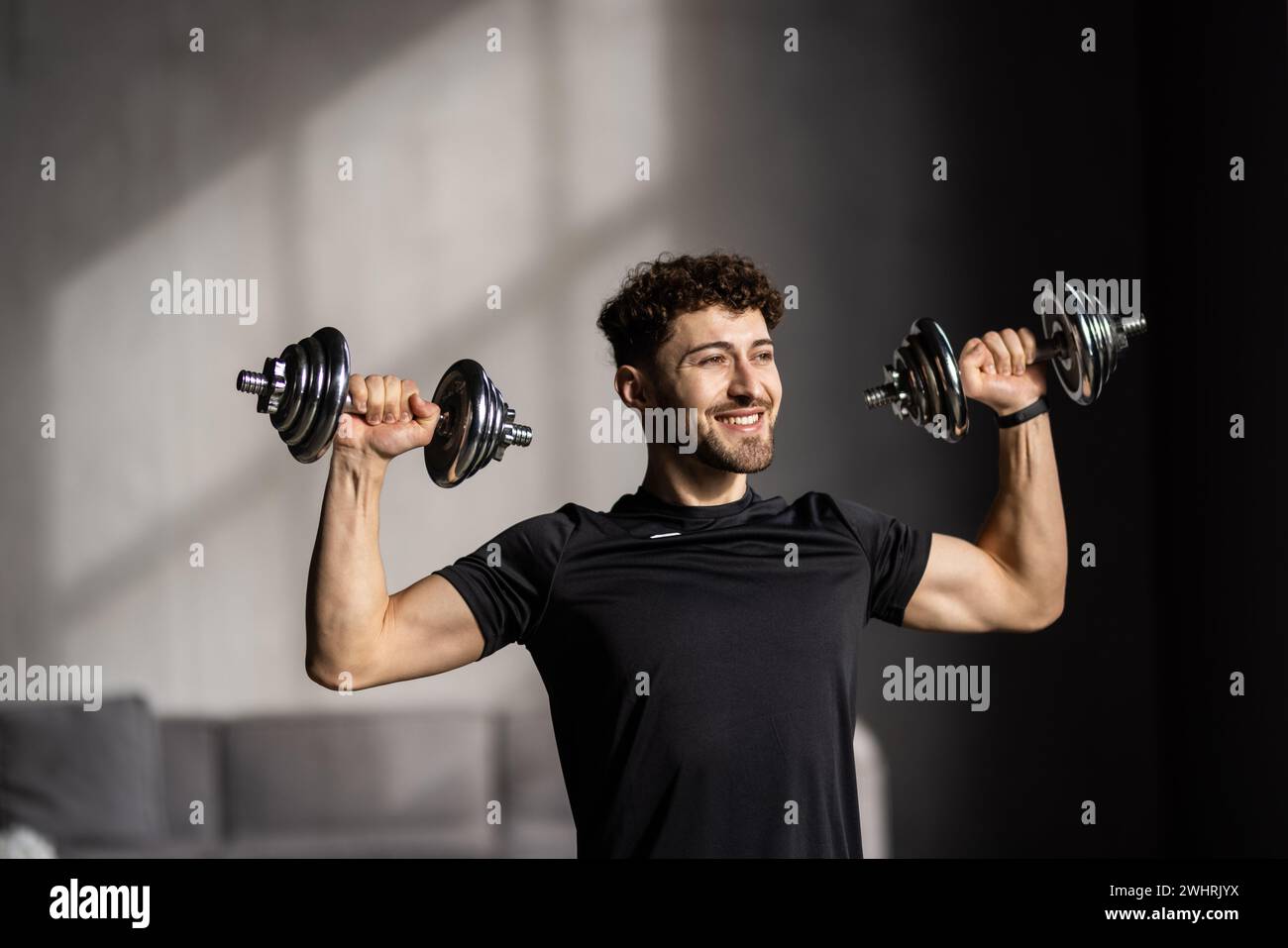 Closeup of handsome middle-aged man doing dumbbell workout at home, working on arms strength, looking at his biceps, copy space. Athletic man lifting Stock Photo