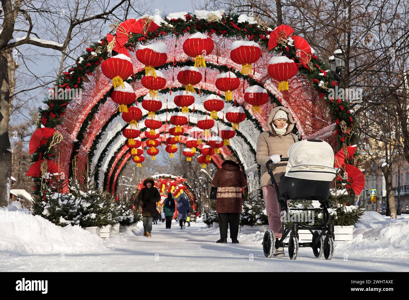 People walking on background of Chinese New Year decorations, woman with pram in foreground. Festive alley decorated with red paper lanterns in winter Stock Photo