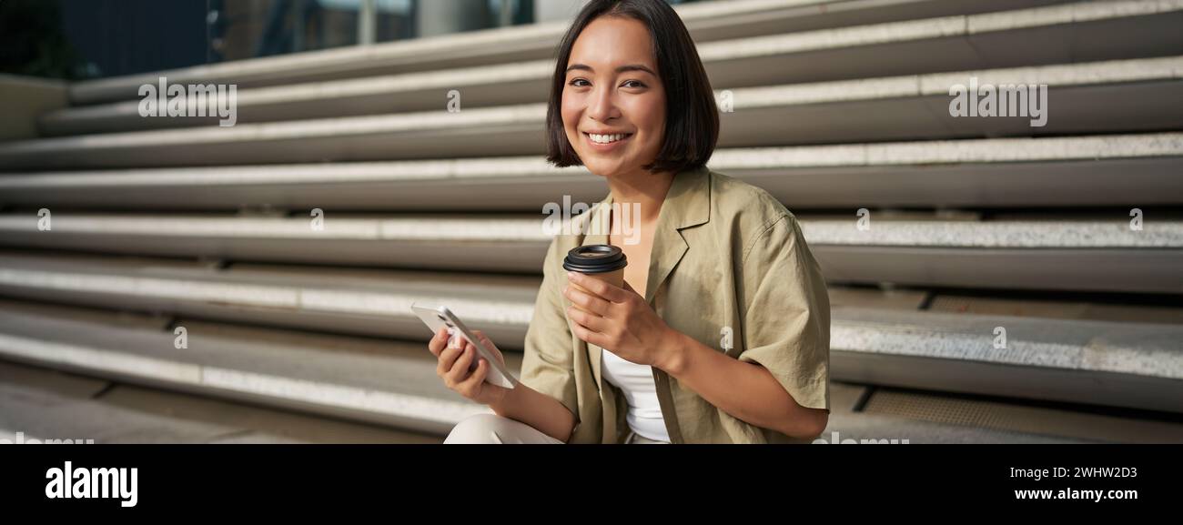 Portrait of asian woman with smartphone, drinks coffee and watches videos on mobile phone. Girl with telephone sits on stairs ou Stock Photo