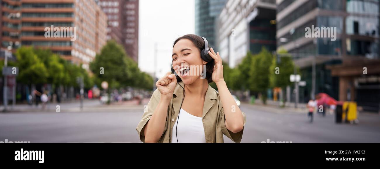 Dancing girl feeling happy in city. Asian woman dancing and listening music in headphones, posing on street Stock Photo
