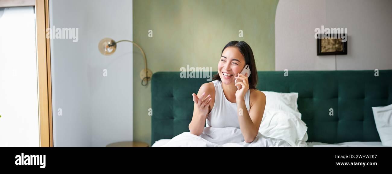 Beautiful smiling asian girl talking on mobile phone, lying in bed with pleased happy face, speaking to someone on telephone Stock Photo