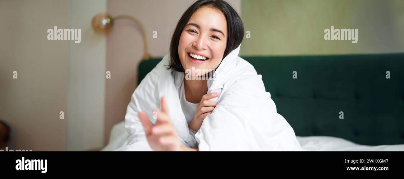 Happy beautiful asian woman covered with blanket, sits on bed in warm duvet, tries to reach camera with a hand, smiling playfull Stock Photo