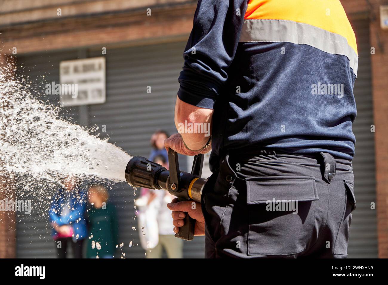 A fireman is seen from behind, handling a fully-engaged hose with a powerful stream of water, likely responding to an emergency Stock Photo