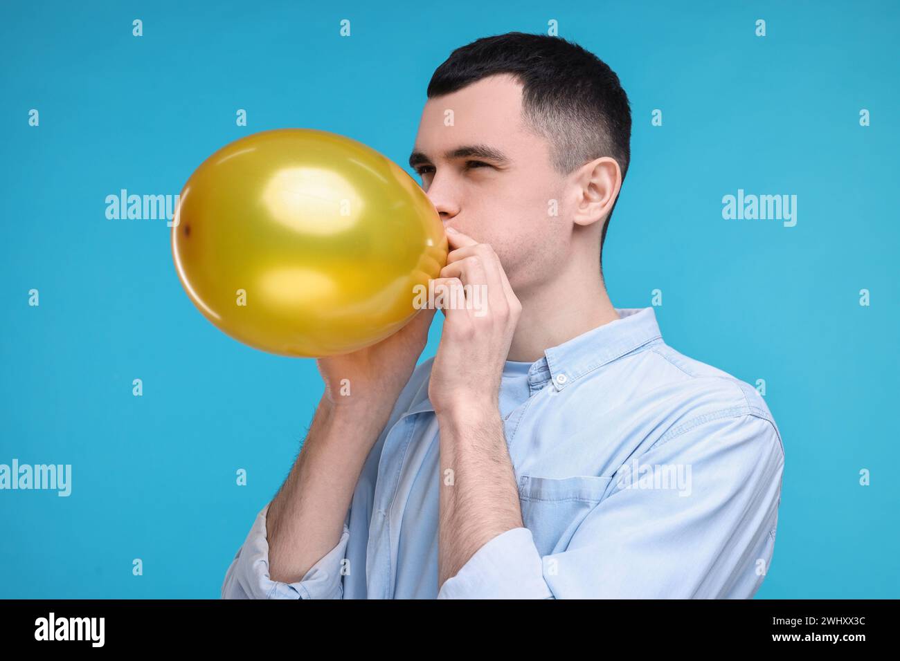 Young man inflating golden balloon on light blue background Stock Photo ...
