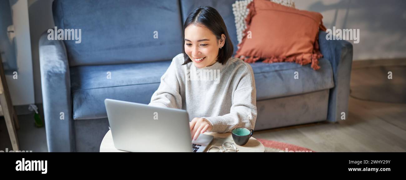 Beautiful asian girl works from home, studying with laptop, sitting in living room, being on remote, freelancing online Stock Photo