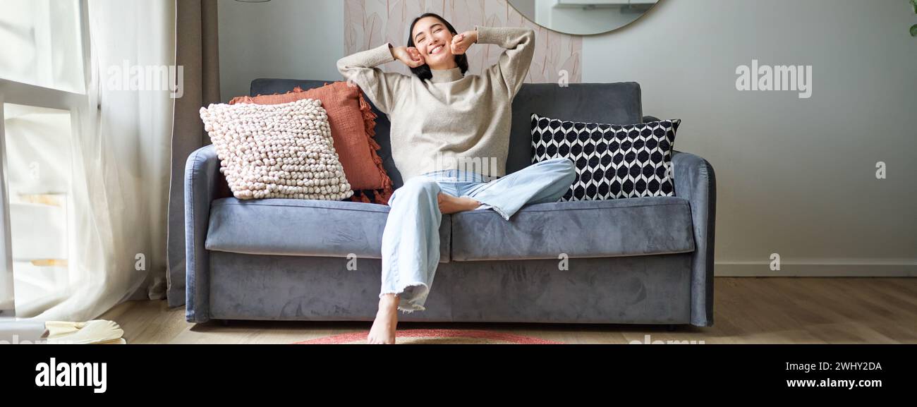 Portrait of happy asian woman feeling lazy, stretching on sofa and smiling pleased, relaxing at home, resting from work Stock Photo
