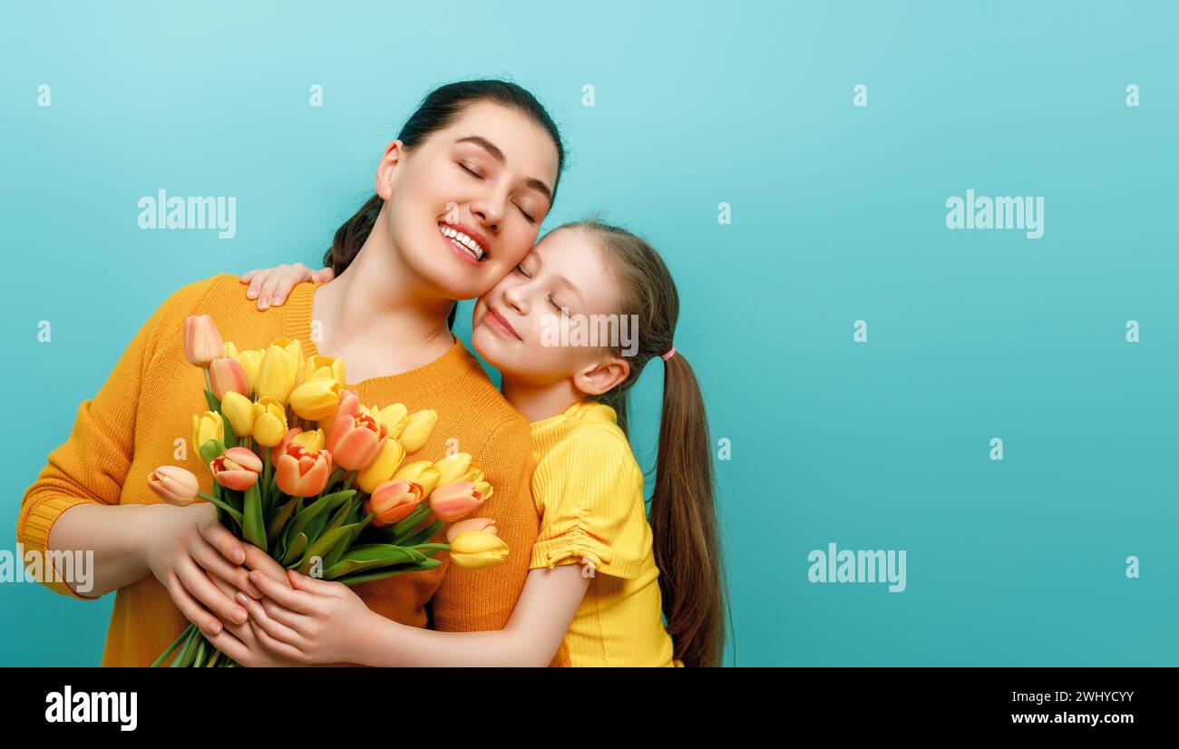 Happy women's day. Child is congratulating mom and giving her yellow flowers. Mum and girl smiling on teal background. Family holiday and togetherness Stock Photo