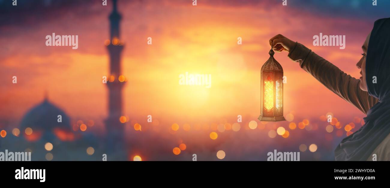 Woman is holding Ornamental Arabic lantern with burning candle glowing at night mosque background. Festive greeting card, invitation for Muslim holy m Stock Photo