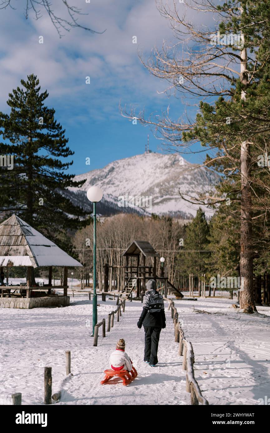 Mom carries a small child on a sled along a snowy path in the park to ...