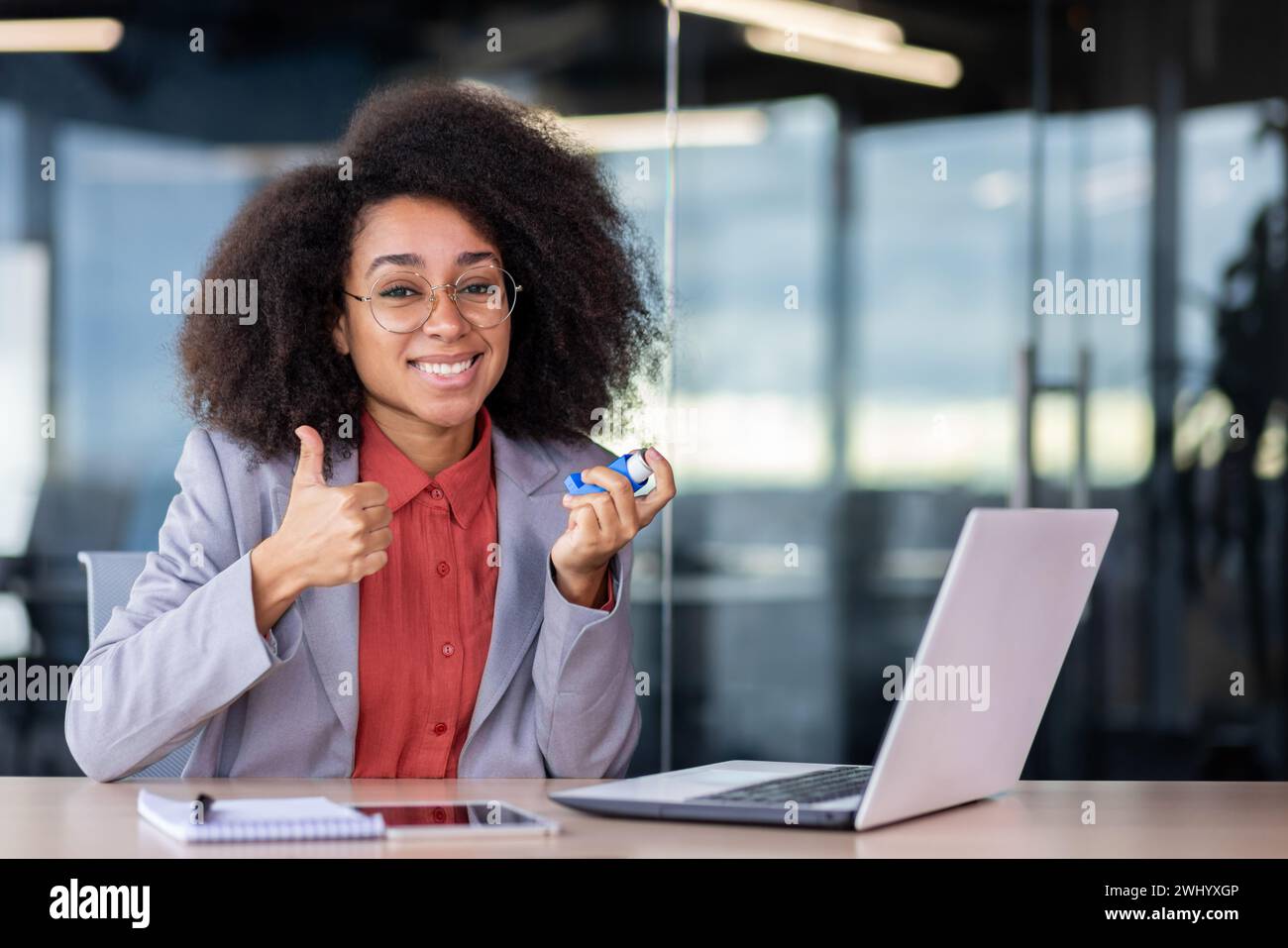 Relieved latino lady in business suit showing thumb um gesture and holding asthmatic aerosol while sitting in cabinet. Smiling administrator feeling better after dealing with bronchial disease attack. Stock Photo