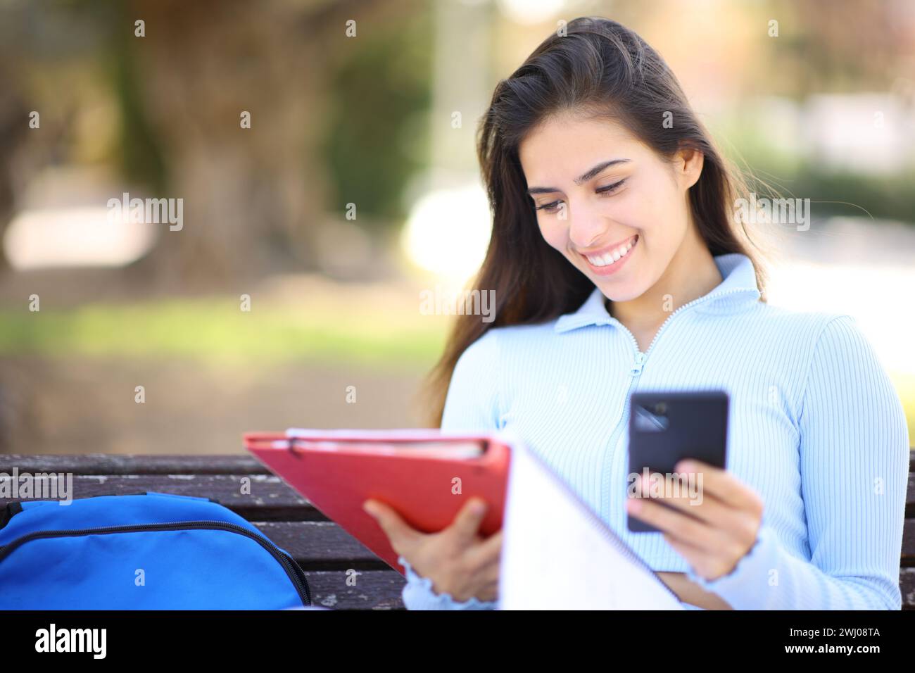 Happy student comparing phone and notes sitting in a park Stock Photo