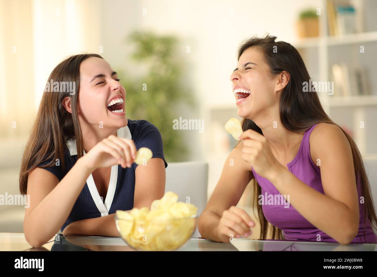 Two happy friends laughing hilariously eating potato chips at home Stock Photo