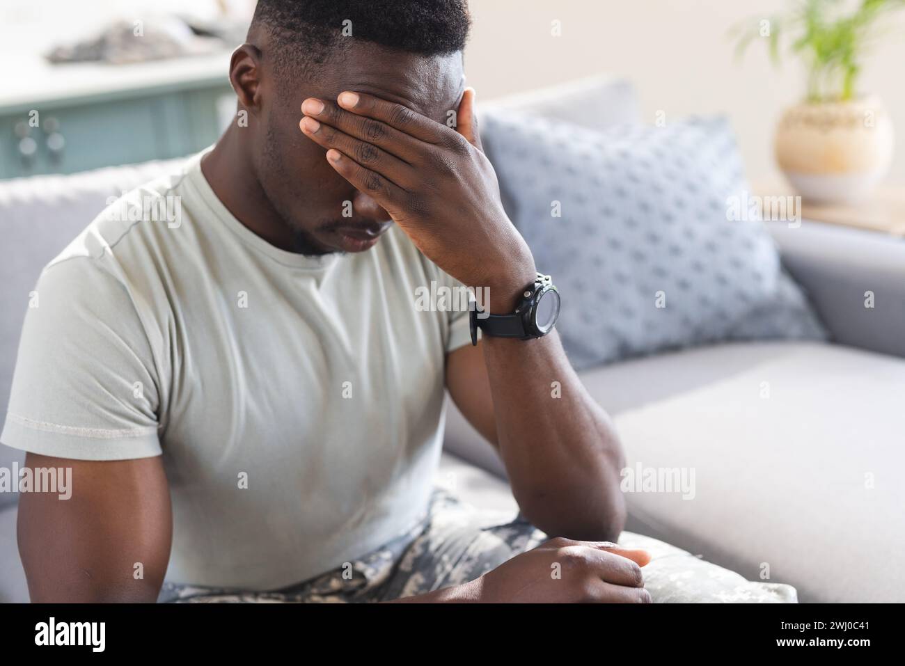 Troubled african american male soldier sitting on couch at home, holding head, copy space Stock Photo