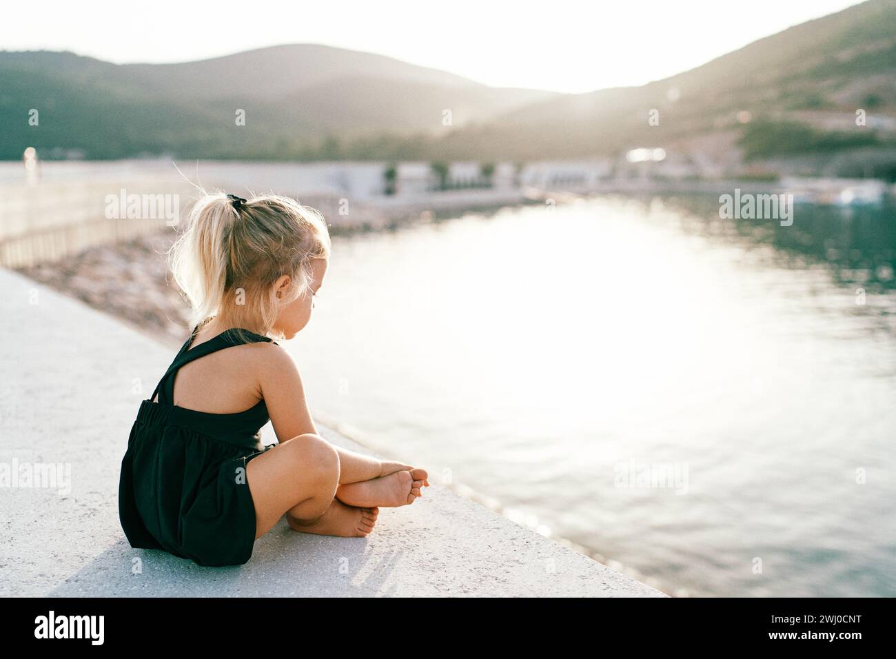 little-barefoot-girl-sits-cross-legged-on-the-pier-and-looks-at-the