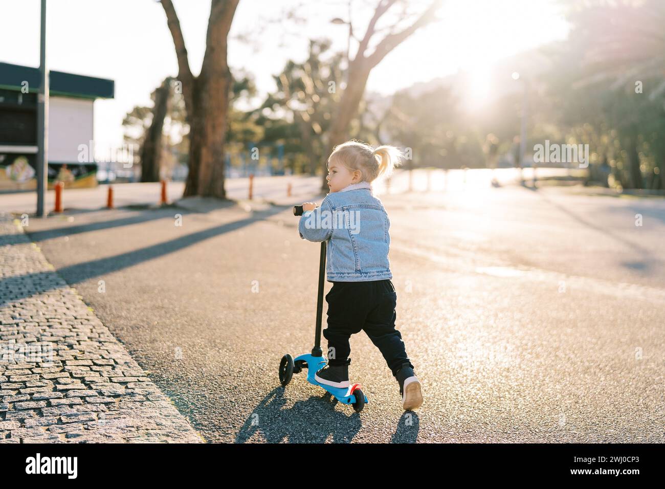 Little girl rides a scooter on an asphalt road and looks away. Back view Stock Photo