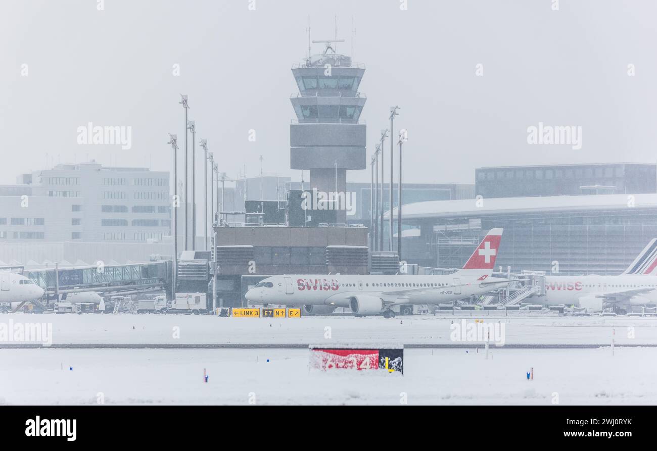 Ein Airbus A220-371 von Swiss International Airlines steht bei starkem Schneefall vor dem Terminal B und dem Kontrollturm des Flughafen Zürich. Regist Stock Photo