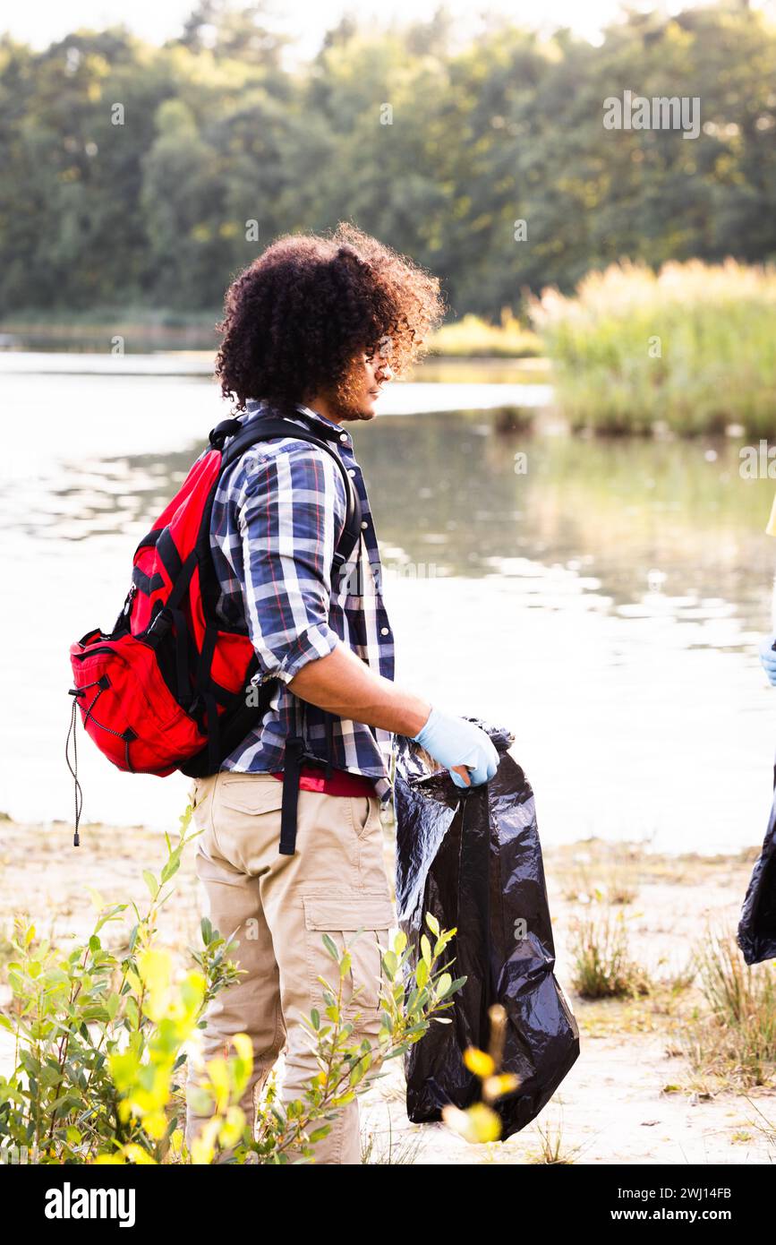 This image portrays a conscientious African American individual with curly hair, taking part in an environmental cleanup. They are dressed in a plaid shirt and khaki pants, geared with gloves, and holding a black trash bag, ready to collect litter. The red backpack suggests they are prepared and organized, possibly a part of a larger conservation effort. The setting is a peaceful lakeside with lush greenery in the background, which emphasizes the importance of the individual's actions in preserving the natural beauty of the area. Eco-conscious Individual Cleaning Lakeside Area. High quality ph Stock Photo