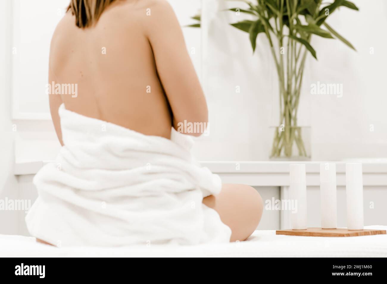 Woman seated on a spa bed, back facing, covered with a towel, beside three beauty products in an aesthetic center, evoking a serene spa ambiance Stock Photo