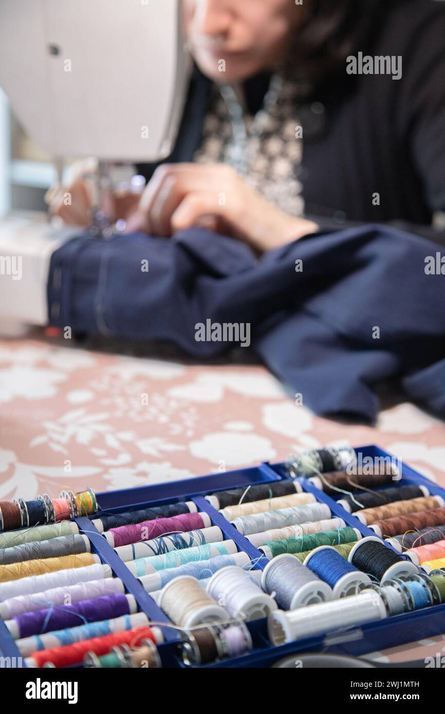 Work table with different sewing utensils and a blurred background with a person sewing with a machine. Stock Photo