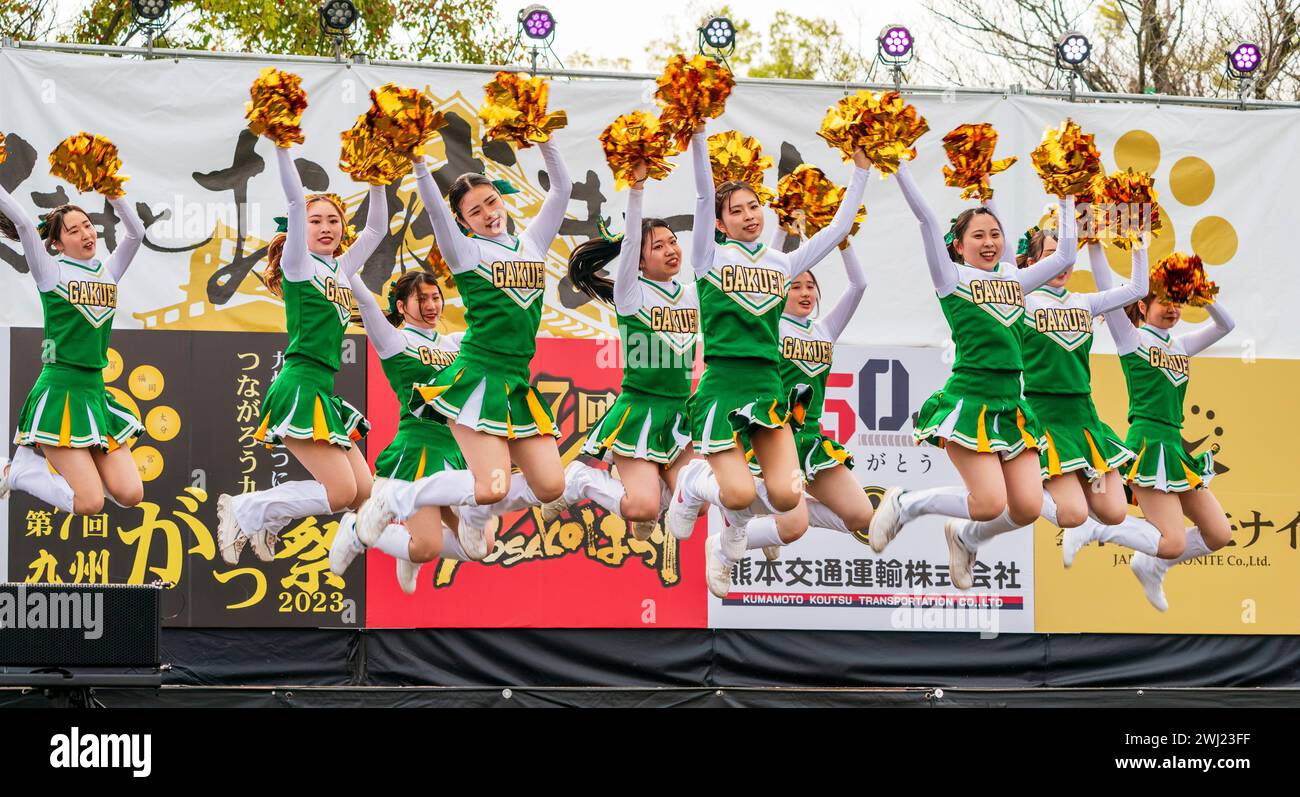 Japanese teenage women cheerleader Yosakoi dance team in green costumes dancing on stage holding gold glittery pom poms. All in mid air after jumping. Stock Photo