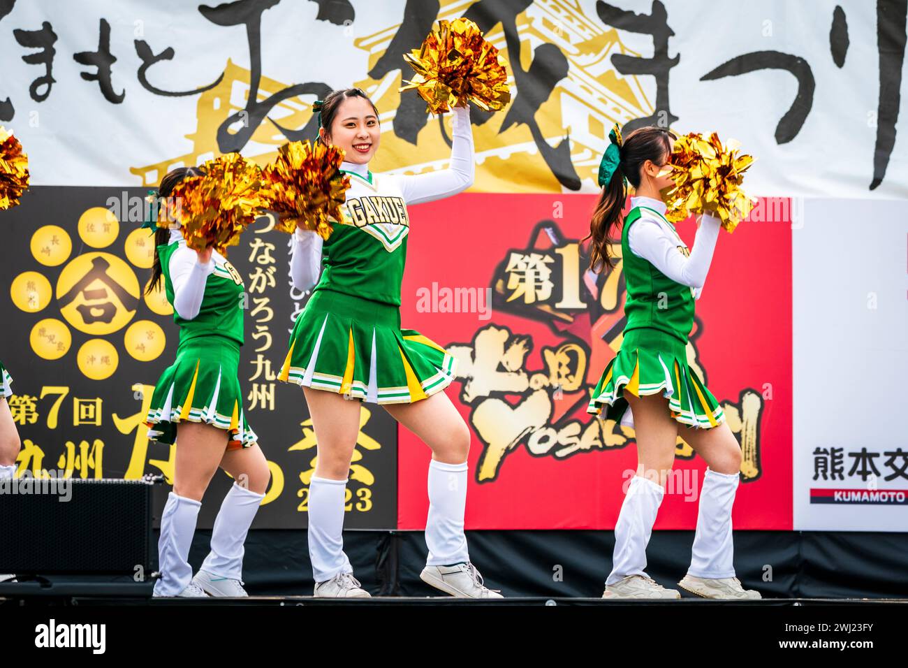 Japanese teenage women cheerleader Yosakoi dance team in green costumes dancing on stage holding gold glittery pom poms, one woman turned to look. Stock Photo