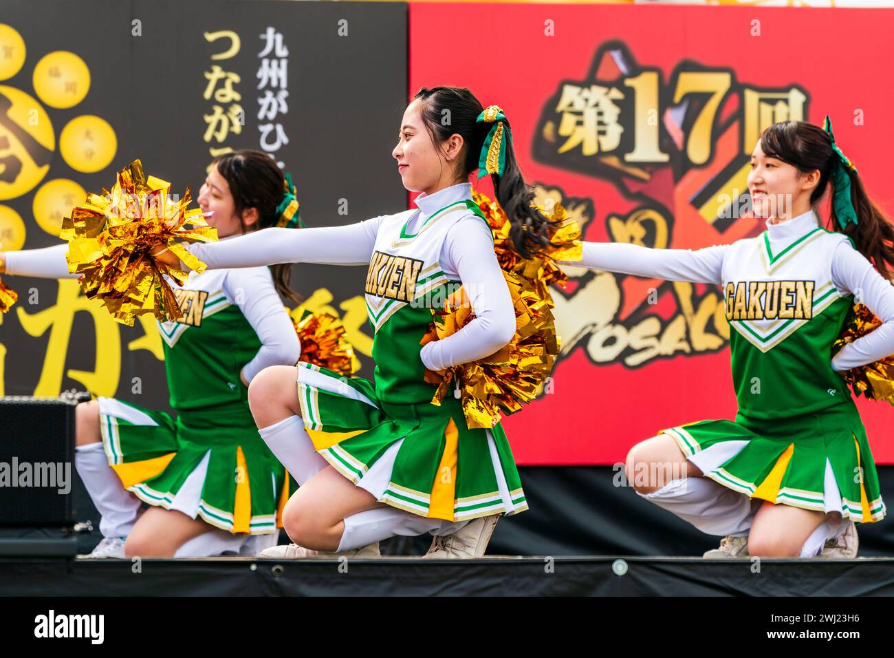 Japanese teenage women cheerleader Yosakoi dance team in green costumes dancing on stage holding gold glittery pom poms. All kneeling on stage. Stock Photo