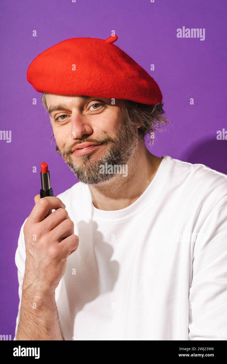 Funny middle aged man wearing red beret is holding lipstick in his hand against purple background Stock Photo