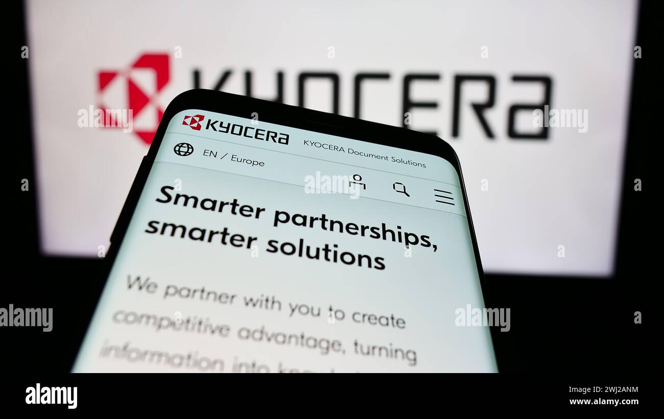 Mobile phone with website of Japanese electronics company Kyocera Corporation in front of business logo. Focus on top-left of phone display. Stock Photo