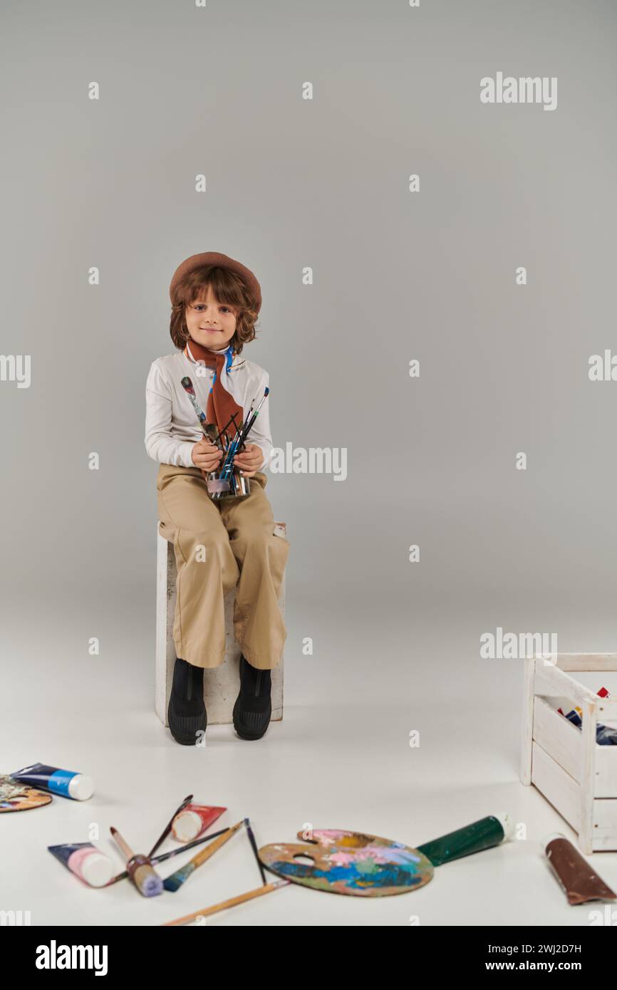 young artist holding glass jar with paint brushes on grey, boy in beret and scarf sitting on chair Stock Photo