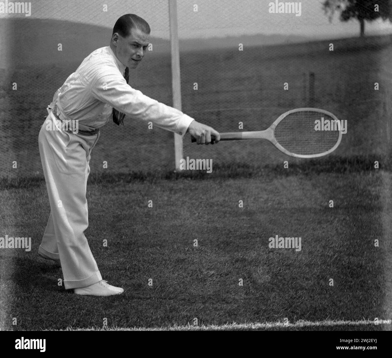 1920s, historical, outside on a grass tennis court, a gentleman tennis player in the stylish sporting dress of the era - long flannel trousers, long sleeve shirt & tie - playing a backhand holding a traditional wooden racquet, England, UK. Stock Photo