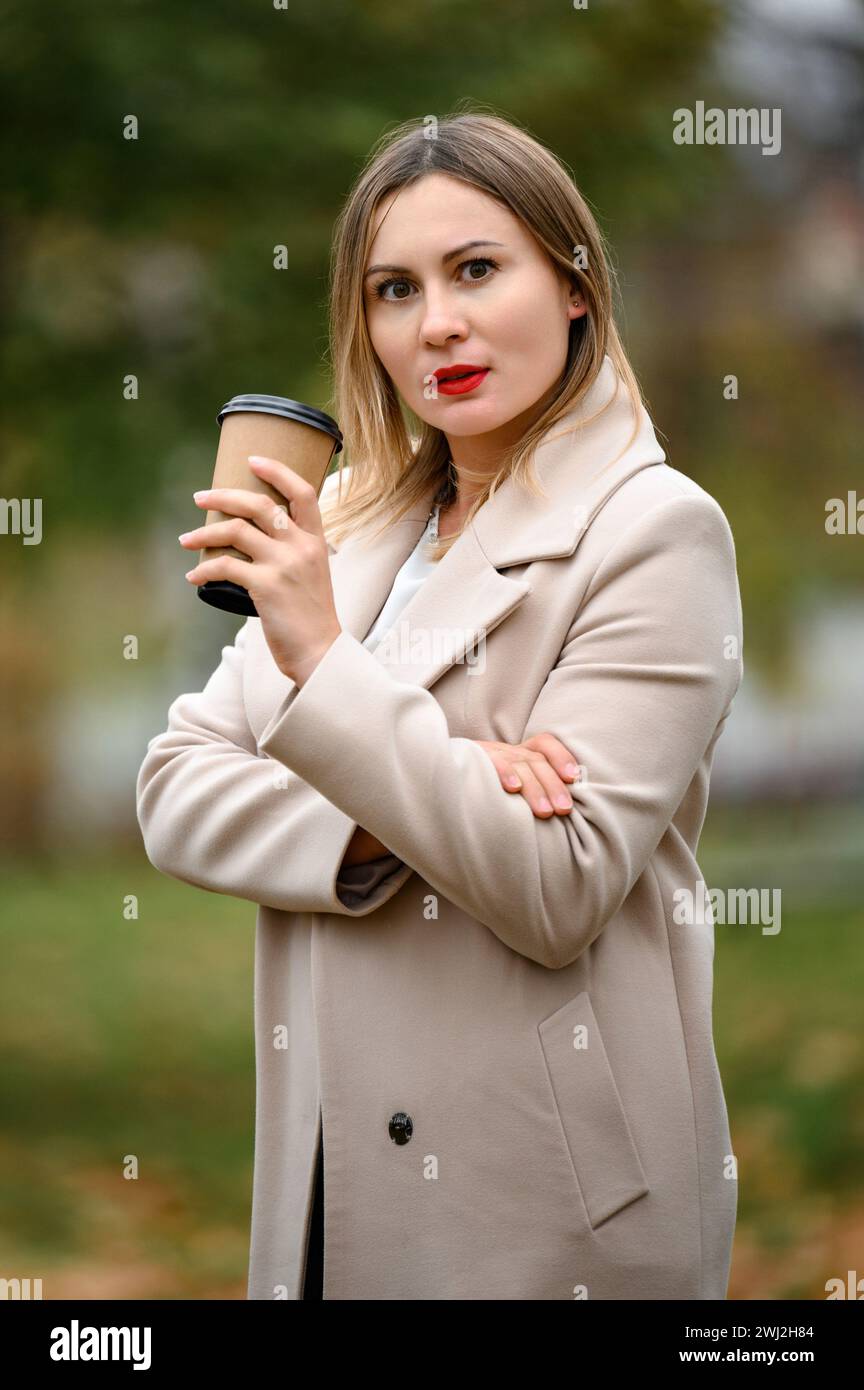A young girl in a long autumn coat drinks coffee in an autumn park, portrait photo of a girl in the park. Stock Photo