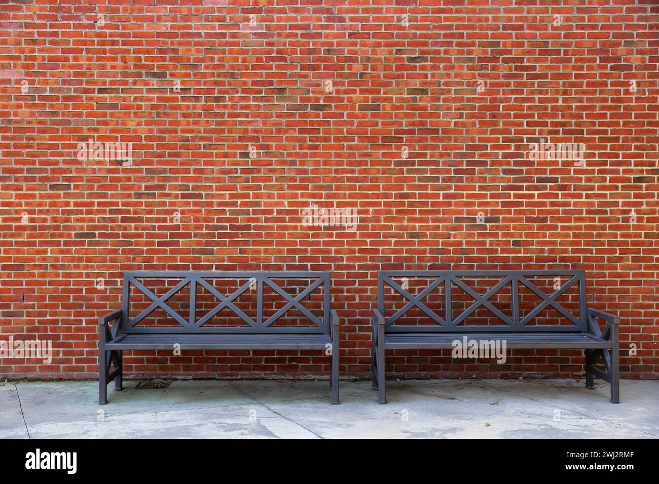 Two iron black benches sit against a brick exterior wall of a building providing copy space and background. Stock Photo
