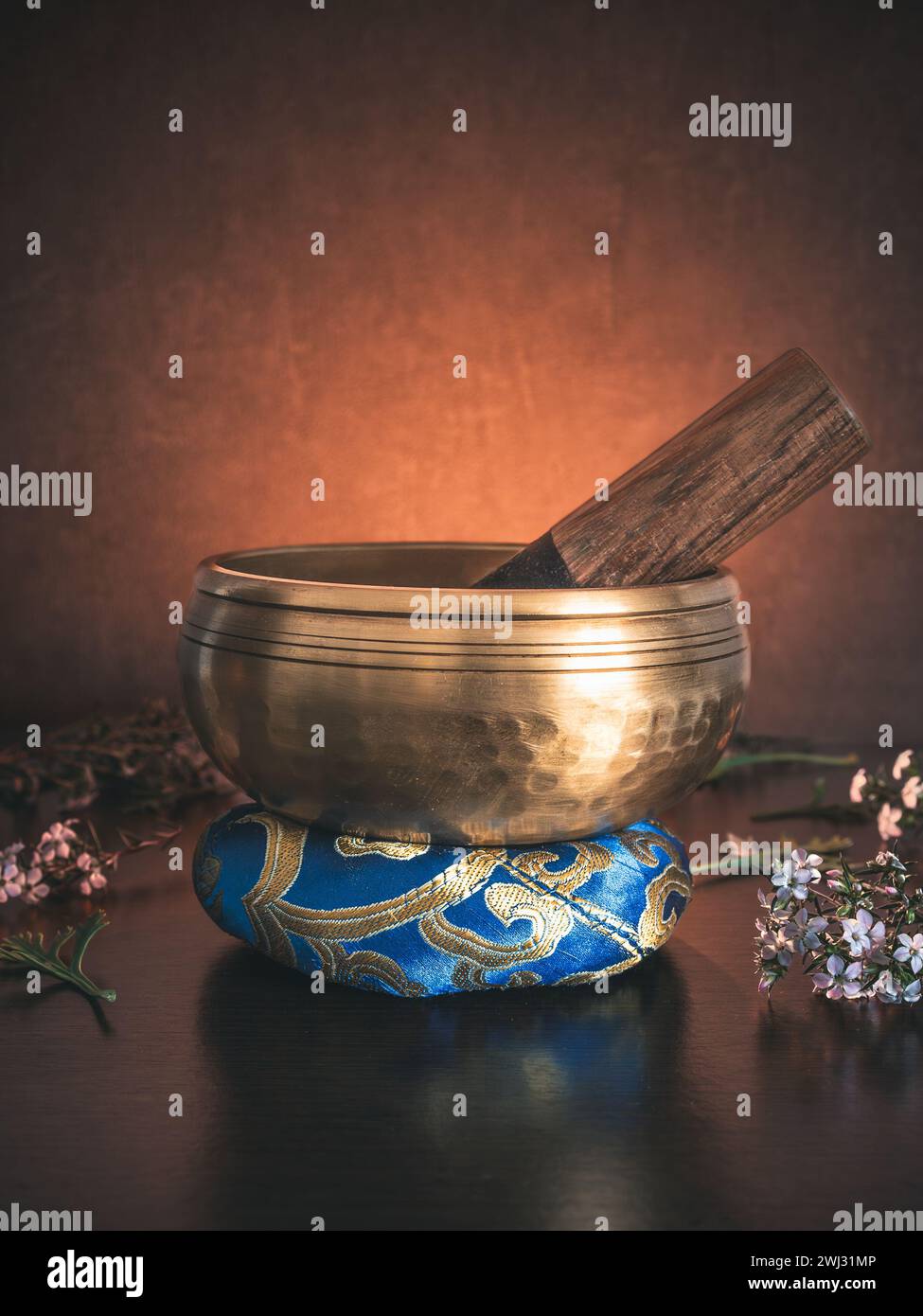 A Tibetan singing bowl sits on a ring cushion amid leaves and flowers, bathed in golden backlight against a dark background Stock Photo