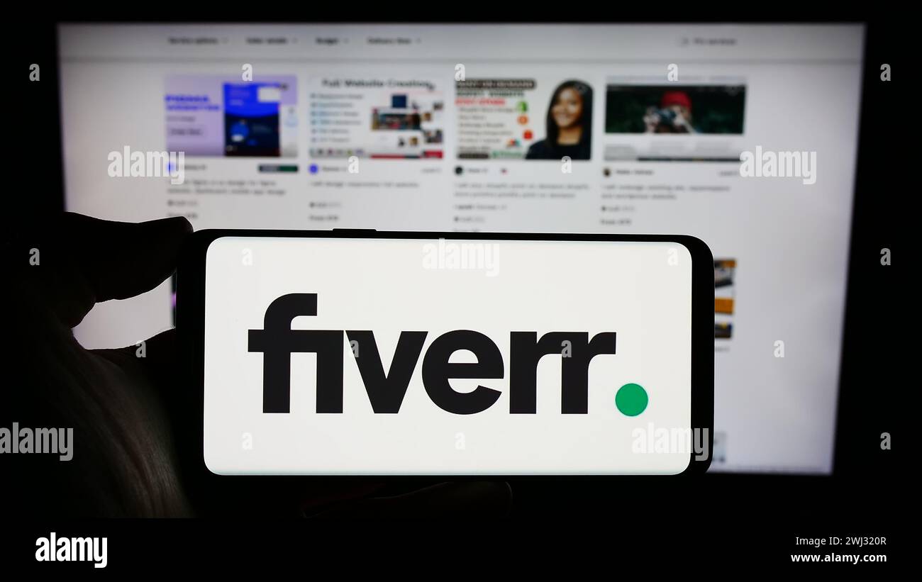 Person holding cellphone with logo of freelance platform company Fiverr International Ltd. in front of business webpage. Focus on phone display. Stock Photo