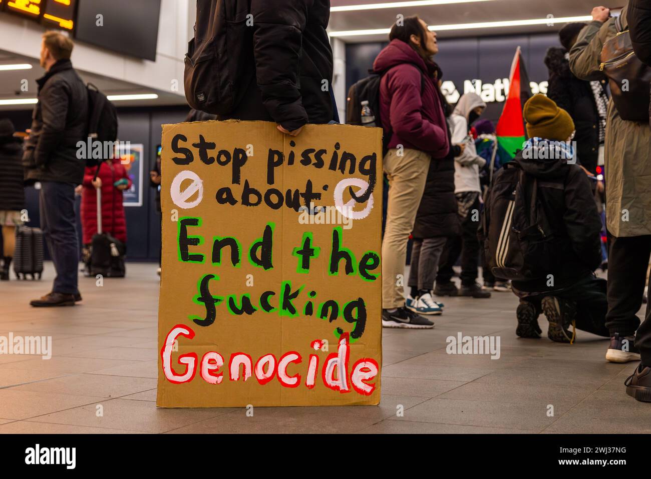 Leeds, UK. 12 FEB, 2024. A protestor besides a sign that reads 'Stop pissing about end the fucking genocide' as part of a pro palestine protest within Leeds station. Credit Milo Chandler/Alamy Live News Stock Photo