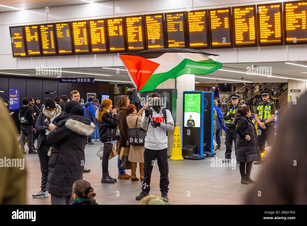 Leeds, UK. 12 FEB, 2024. A masked pro palestine protestor waves palestinian flag infront within the main throughfare for Leeds train station. Credit Milo Chandler/Alamy Live News Stock Photo