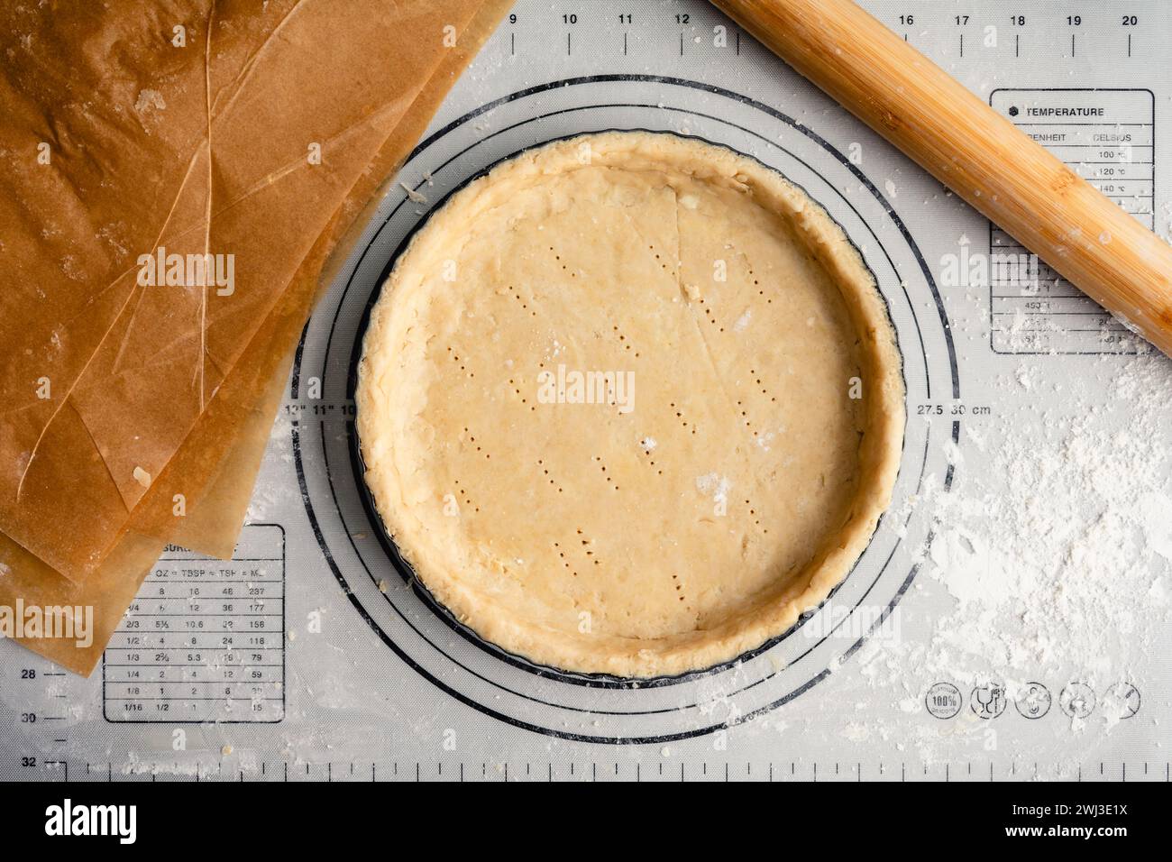 Unbaked Pastry Dough in a Tart Pan: Raw pie crust in a fluted pan on a floured work surface with a French rolling pin and unbleached parchment paper Stock Photo