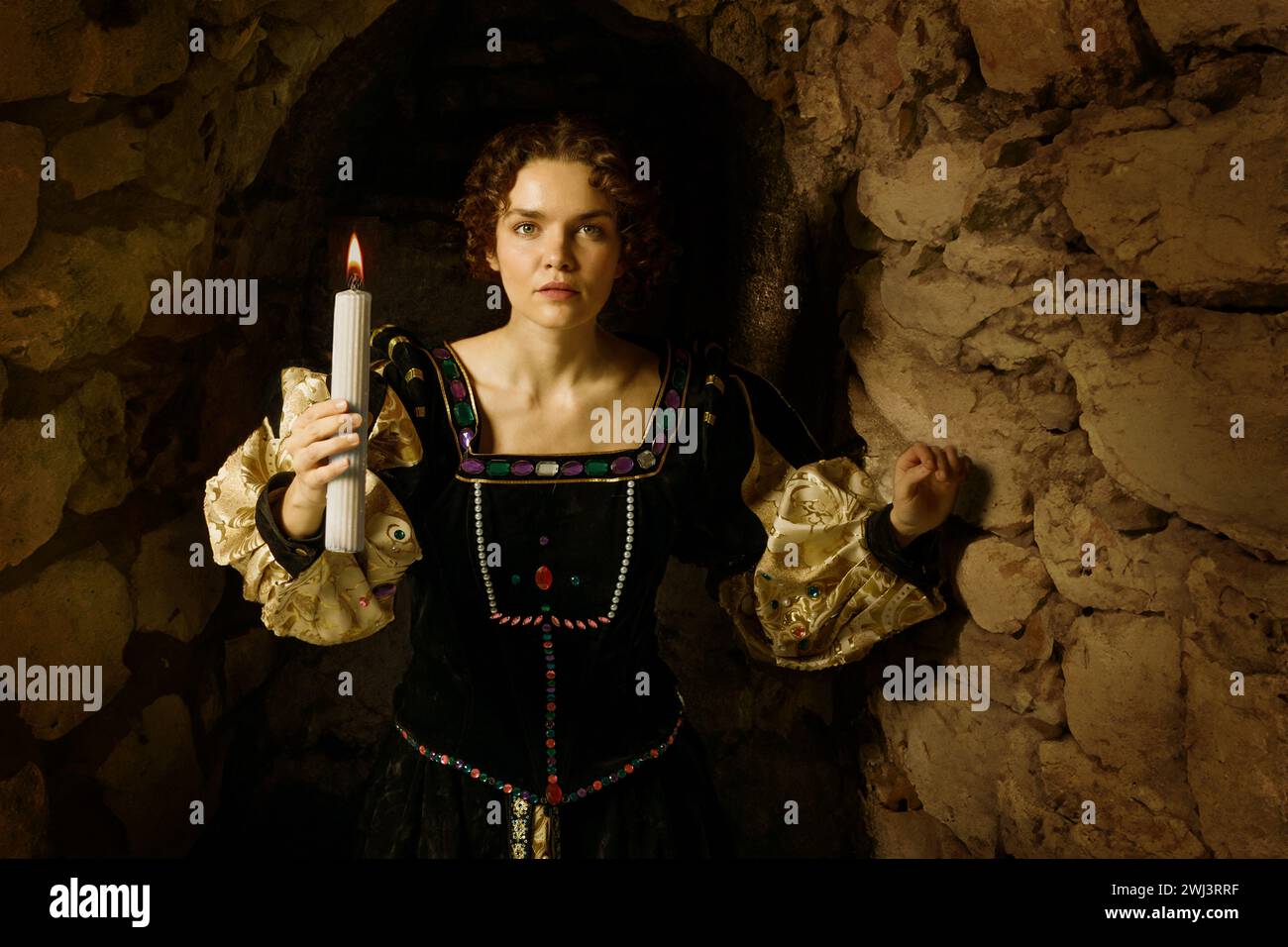 Woman dressed in a medieval dress holding a candle and looking for something on the dark dungeon in the castle. Stock Photo