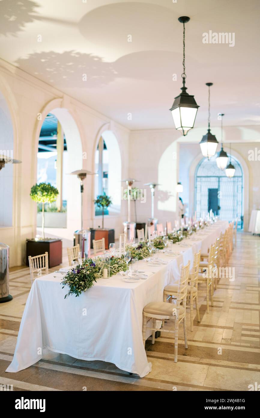 Festive long table with bouquets of flowers in a large hall with glowing chain lamps on the ceiling Stock Photo
