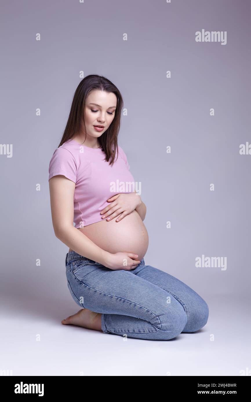 Young pretty pregnant woman in pink t-shirt and jeans sit on the floor on gray background. Female with belly exposed. Stock Photo