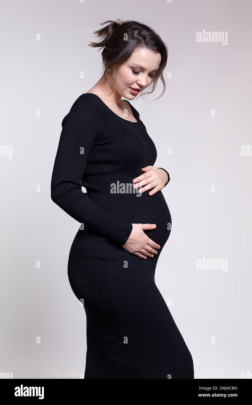 Portrait of young pregnant female in black dress with hands near pregnant belly. Stock Photo