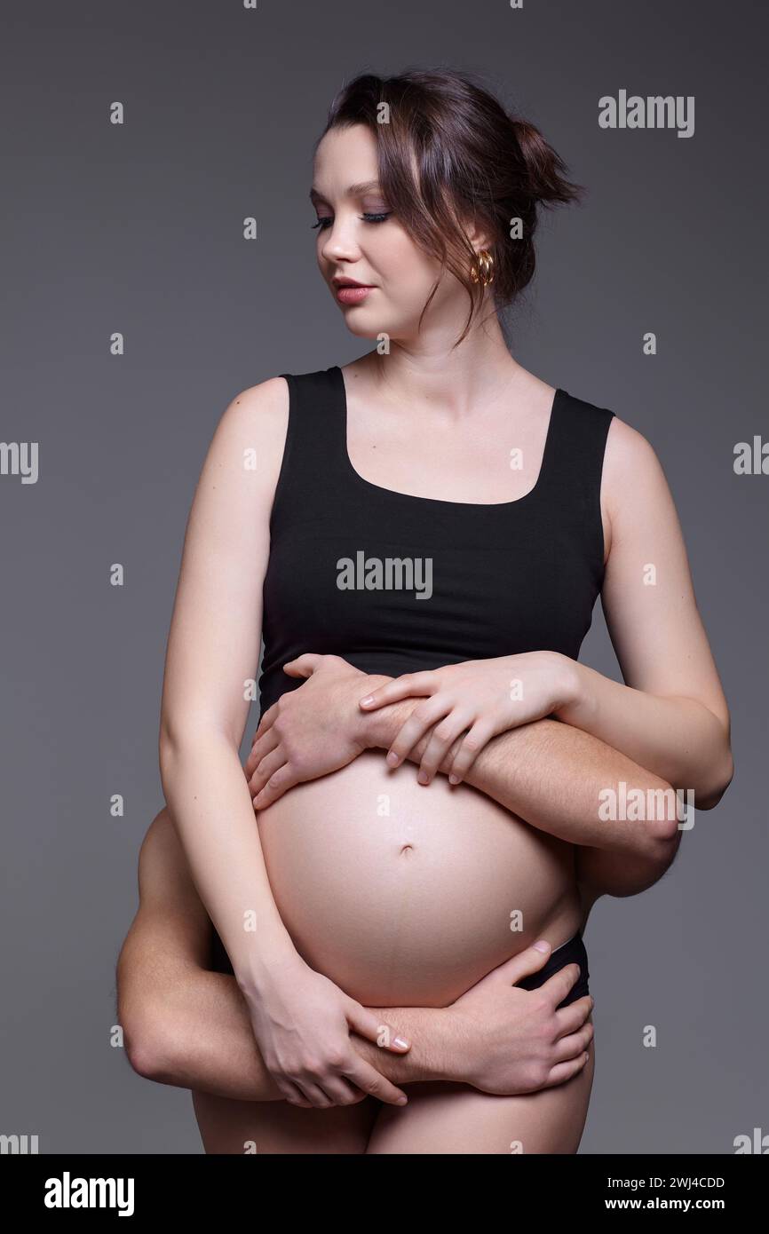 The man is behind female and hugs her pregnant exposed belly. Stock Photo