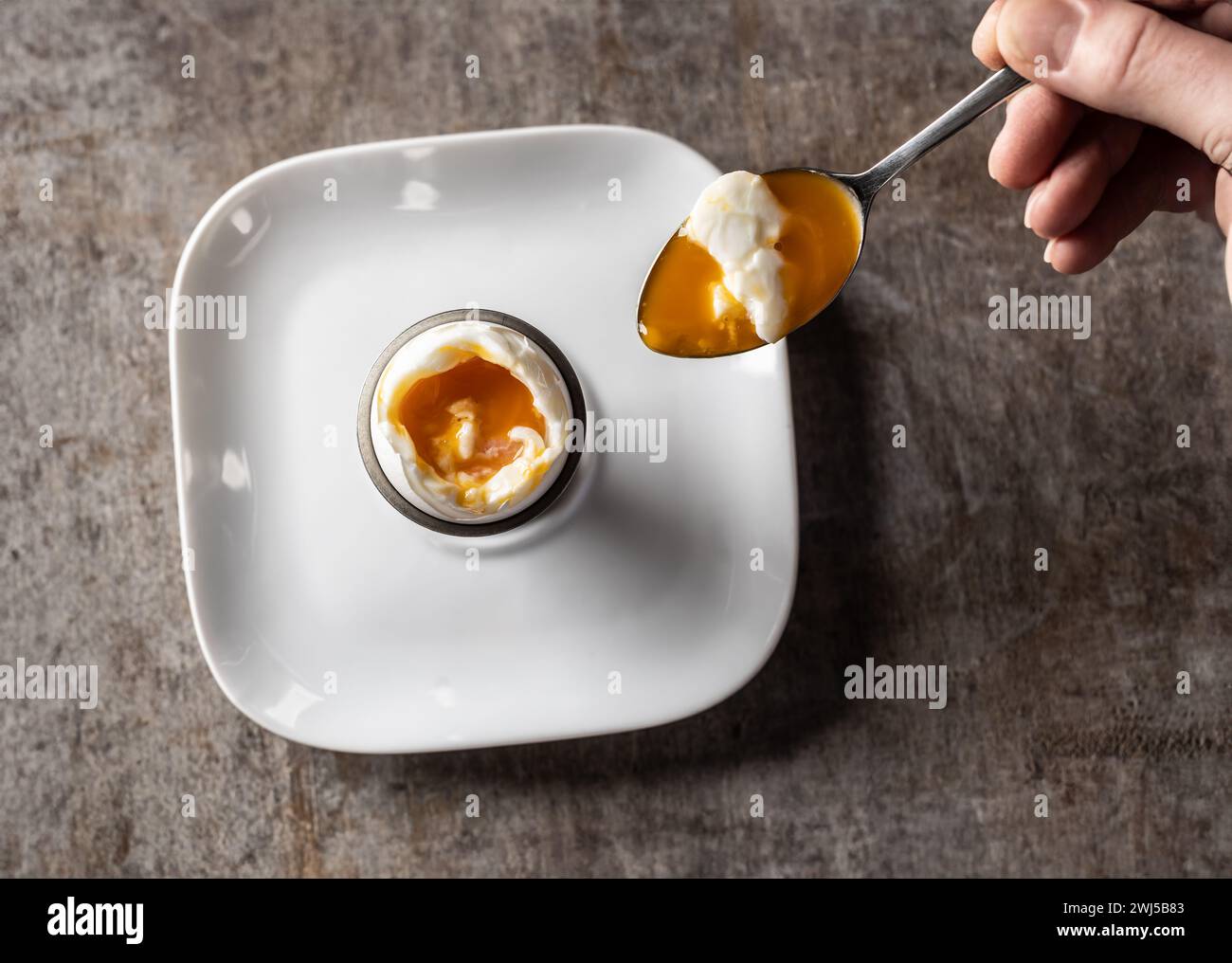 A woman's hand with a spoon scoops a soft-boiled egg from the shell - Top of view. Stock Photo